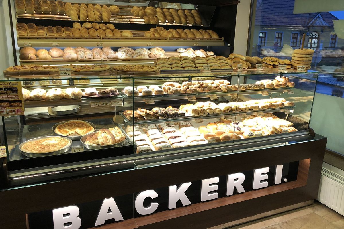 Verkaufstheke der Bäckerei Gazi: Regale mit Brot und Gebäck, darunter eine Vitrine mit Pizza, Pizzaschnitten und Gebäck, beschriftet mit „Bäckerei“